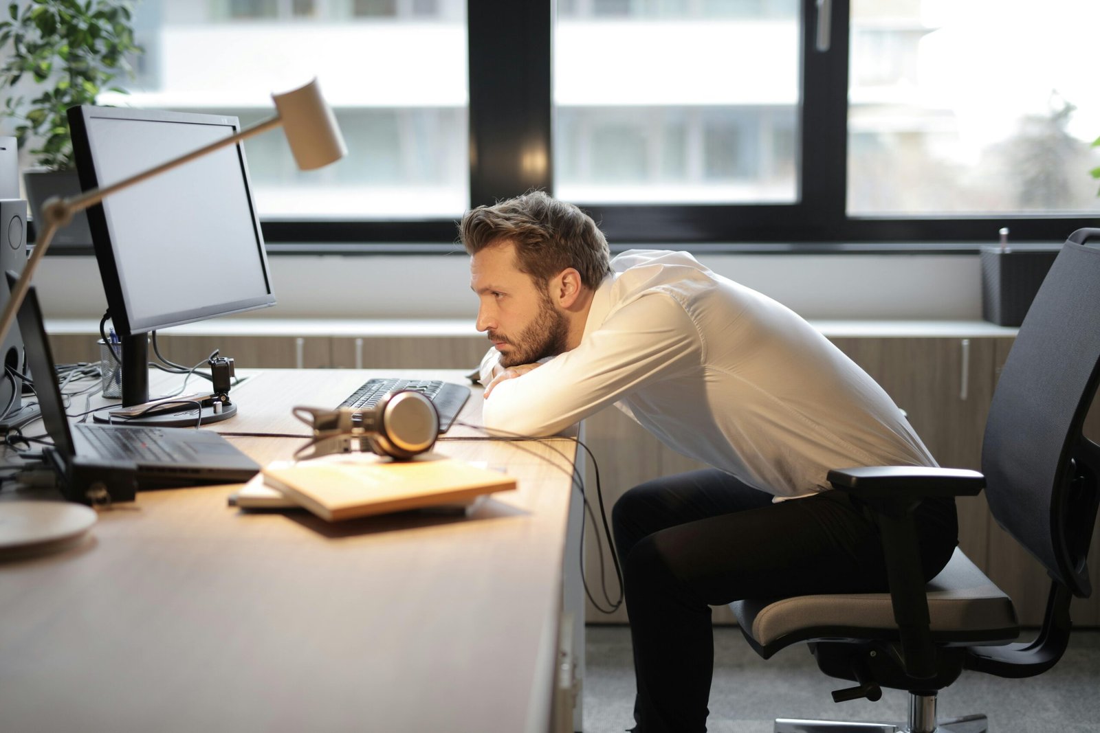 A tired man leaning on his desk in a modern office, looking at the computer screen.